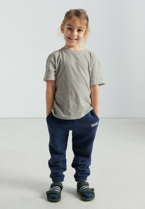 Ragazza giovane e sorridente in piedi con le mani nelle tasche dei pantaloni della tuta blu navy, indossa una maglietta grigia, calzini a righe e sandali verdi su uno sfondo chiaro.