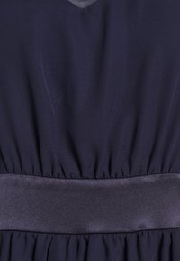 Marindress med en texturerad tyg, V-ringning och en hopkrupen midja med ett satinliknande midjeband. Betoning på veck och släta accenter.