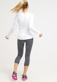 Camiseta atlética de manga larga blanca, combinada con leggins capri de patrón ondulado en blanco y negro y zapatillas de correr rosas. Textura suave, diseño ajustado.