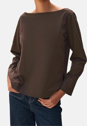 Osoba ubrana w ciemnobrązową bluzkę z długim rękawem i dekoltem typu łódka oraz ciemnoniebieskie dżinsy, z rękami opartymi na biodrach.