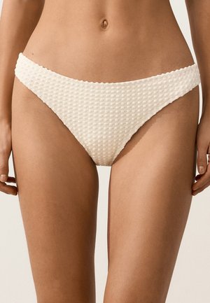 Vrouw draagt witte bikinionderkant met textuur, staande met handen ontspannen langs het lichaam, zichtbaar vanaf de taille tot de bovenkant van de dijen.