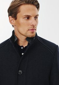 Granatowy wełniany płaszcz z wysokim kołnierzem i szarym akcentem przy dekolcie, z pojedynczym zapięciem na guzik i subtelnymi detalami w stębnowaniu.