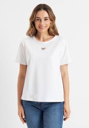Kobieta uśmiechająca się, ubrana w białą koszulkę z krótkim rękawem z małym wzorem serca na środku oraz niebieskie dżinsy na jednolitym tle.