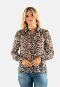Blusa trasparente nera e beige con un motivo astratto a foglia, chiusura con bottoni frontale, maniche lunghe e colletto classico. Abbinata a jeans blu.