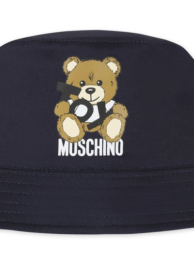 Cappellino blu navy con un design di un orso cartoon. L'orso tiene in mano una palla, con 'TOY' e 'MUSCHINO' stampati in bianco sottostante. Texture liscia.