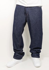 Dark blue denim jeans with a relaxed fit, featuring white stitching and pockets, paired with white sneakers.