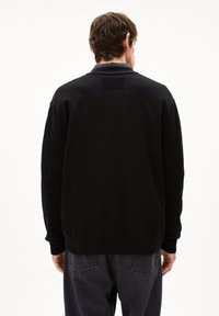 Schwarzer Strickpullover mit lockerem Schnitt, Rundhalsausschnitt und gerippten Bündchen. Mit einem strukturierten Patch-Detail auf dem oberen Rücken. Getragen mit grauen Jeans.