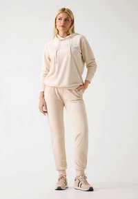 Bež pulover s kapuco z logotipom »GUESS«, kombiniran s hlačami v istem stilu. Gladek material, sproščen kroj in stiskani manšeti. Superge s črnimi poudarki.