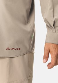 Camicia beige a maniche lunghe con tessuto texturizzato, caratterizzata da un logo ricamato color bordeaux e cuciture a contrasto, mostrata con una mano all'estremità della manica.