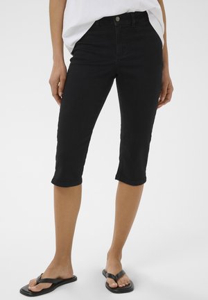 Donna con pantaloni neri aderenti fino al ginocchio, top bianco e infradito nere, in piedi contro uno sfondo chiaro e semplice.