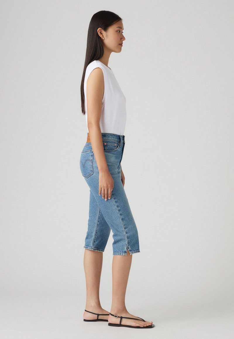 Haut sans manches blanc, jean bleu taille mi-hauteur avec des bords effilochés et des fentes sur les côtés, associé à des sandales noires. Tenue simple et décontractée.