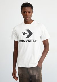 T-shirt bianca in cotone con un grande logo nero stampato con un design di stella e freccia, abbinata a pantaloni cargo marroni.