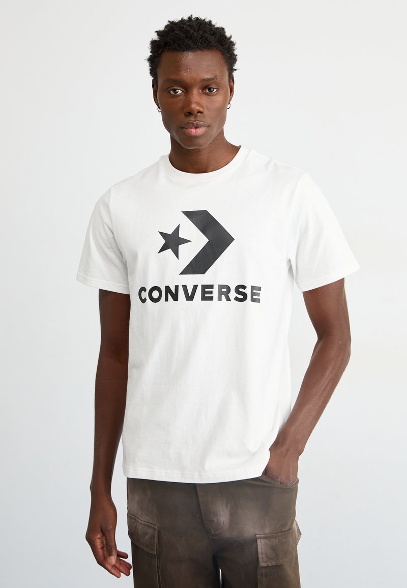 T-shirt bianca in cotone con un grande logo nero stampato con un design di stella e freccia, abbinata a pantaloni cargo marroni.