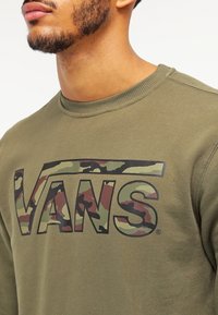 Sudadera verde oliva con cuello redondo. Presenta un gran logo "VANS" de camuflaje multicolor en el pecho. Textura suave, corte casual.