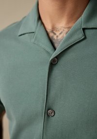 Camisa verde con cuello, fabricada con un tejido texturizado y con cierre de botones. Vista en primer plano que muestra los botones y el detalle del cuello.