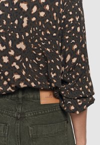 Leopardenmuster-Bluse mit hochgeschlagenen Ärmeln, in hoch sitzende olivgrüne Hosen gesteckt, die ein Leder-Label auf der Taille haben.