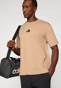 T-shirt en coton beige avec col rond et manches courtes, arborant un logo Adidas noir sur la poitrine, accompagné d'un sac de sport noir.