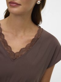 Γυναίκα που φοράει κα φέ μπλούζα με V λαιμόκοψη και δαντέλα, με ένα μεγάλο στρογγυλό μαργαριταρένιο σκουλαρίκι στο εμφανές αυτί.