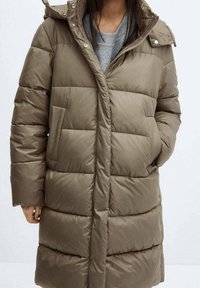 Manteau matelassé vert olive avec fermeture éclair et boutons-pression, présentant des coutures horizontales, des poches latérales et une capuche. Texture lisse et rembourrée.