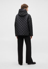 Melnā izšūtā jaka ar kapuci, kurai ir dimanta raksts. Apģērbs ir brīva piegriezuma un tas ir kombinēts ar melna platā stila biksēm.