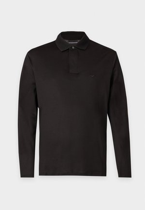 Junger Mann, der ein schwarzes langärmeliges Polohemd, lockere schwarze Hosen und weiße Turnschuhe vor einem schlichten hellen Hintergrund trägt.