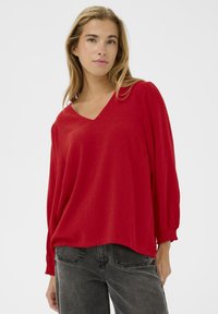 Rode V-hals blouse met lange, opgeblazen mouwen, gemaakt van lichtgewicht stof. Heeft een losse pasvorm en een textuur op het oppervlak. Gekleed met donkere jeans.