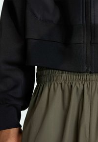 Crni skraćeni dukserica sa zatvaračem i maslinasto zelene hlače s elastičnim strukom, koje imaju detalje od nabranog materijala. Vidljive su glatke teksture.