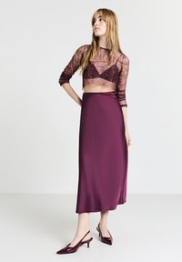 Top corto in pizzo di un profondo color porpora abbinato a una gonna midi in satin dello stesso colore. La gonna ha una trama liscia; le scarpe sono di tipo slingback in una tonalità simile.