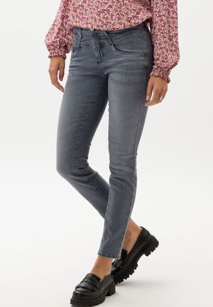 Frau trägt eng anliegende graue Jeans, schwarze Loafers mit Fransen und eine rosa geblümte Langarmbluse, steht mit übergeschlagenem Bein.