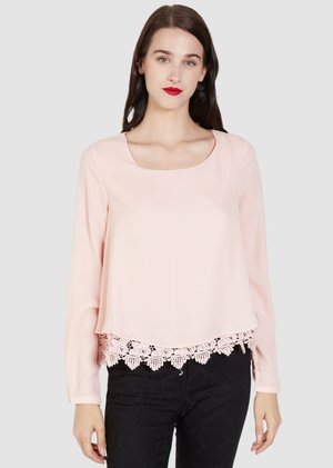 Mujer con cabello largo castaño, vistiendo una blusa rosa pálido de manga larga con dobladillo de encaje y pantalones negros, de pie contra un fondo gris claro.