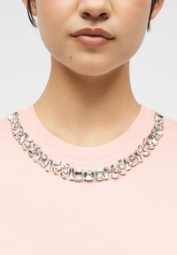 Εγγύς πλάνο ενός ατόμου που φοράει ροζ ανοιχτό τοπ με στρογγυλή λαιμόκοψη διακοσμημένη με διάφορους διαφανούς, πολυεδρικούς πολύτιμους λίθους.