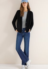 Femme portant un blazer noir, un pull gris, un jean bleu, des baskets argentées et une casquette bleu clair, debout les mains dans les poches devant un fond neutre.