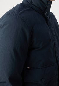 Man in een donkerblauwe gewatteerde jas met een hoge kraag en een drukknoop-flapzak op de borst.