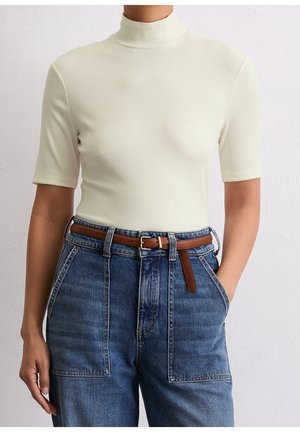 Kobieta ubrana w kremowy, prążkowany top z krótkim rękawem i stójką, włożony do niebieskich dżinsów z wysokim stanem, z przednimi kieszeniami naszywanymi i brązowym paskiem.