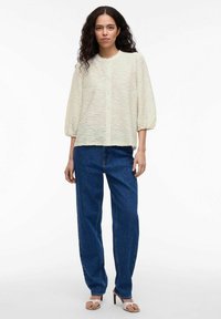 Light beige textured blouse with a round neckline, button closure, and puff sleeves, paired with loose blue jeans and light sandals.