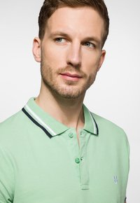 Zielona koszulka polo z fakturowanym materiałem, z zapięciem na dwa guziki i kontrastującymi granatowymi paskami na kołnierzyku. Małe logo na lewej piersi.