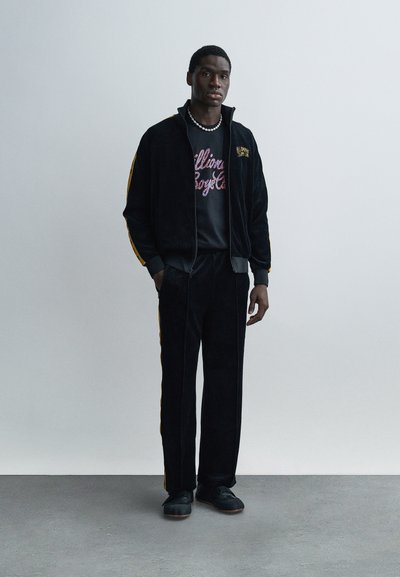 Μαύρο φόρμα βελουτέ με κίτρινες πλευρικές ρίγες, συνδυασμένο με ένα γραφικό μπλουζάκι με πολύχρωμο κείμενο και μαύρα αθλητικά παπούτσια.