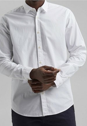 Mand iført en hvid langærmet skjorte med knapper, der justerer manchet med begge hænder mod en ens grå baggrund.