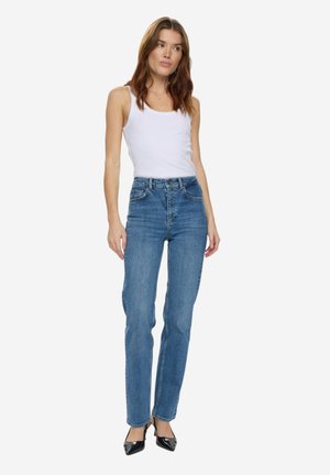 Kvinde stående iført en hvid ærmeløs top, højtaljede blå jeans og sorte spidse hæle foran en ensfarvet baggrund.