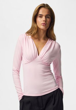 Femme aux longs cheveux châtain clair portant un haut rose clair à manches longues en V et un pantalon noir, debout devant un fond uni.