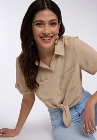 Camicia beige a maniche corte con bottoni, realizzata in un tessuto texture, due tasche frontali e orlo annodato, abbinata a jeans in denim azzurro chiaro.