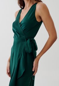Smaragdgrön omslagsklänning i satinmaterial, med V-ringning, breda band och knytning i midjan, samt lager på lager volangdetalj.