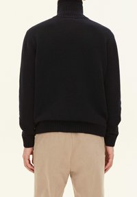 Maglione lavorato a maglia colore navy con un alto colletto a coste, spalle scese e polsini e fondo a coste. Indossato su pantaloni beige.