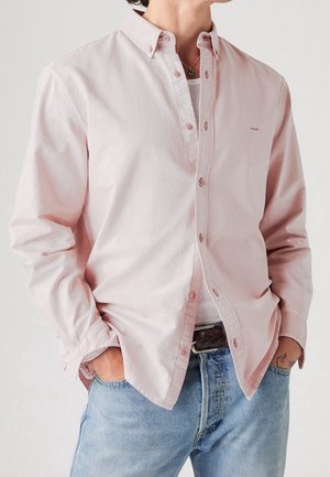 Mężczyzna w jasnoróżowej koszuli na guziki, nałożonej na biały tank top, w połączeniu z niebieskimi dżinsami i czarnym plecionym paskiem, ręce w kieszeniach.