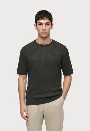 Junger Mann trägt einen dunkelgrünen, strukturierten kurzärmeligen Pullover und hellbeige Hosen, steht mit einer Hand in der Tasche auf einfarbigem Hintergrund.