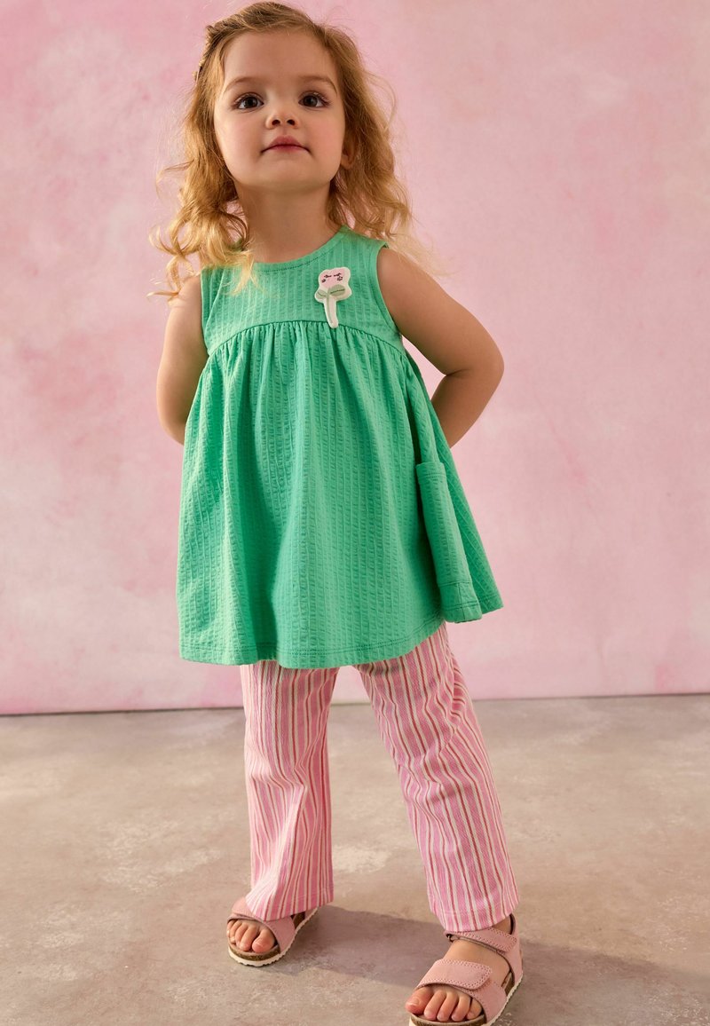 Young girl with curly hair wearing a green sleeveless dress, pink and white striped pants, and pink sandals, standing against a pink background.