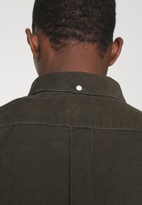 Donkergroene corduroy shirt met een gestructureerde kraag en een witte knop aan de achterkant, met een verticale ribbelstructuur.