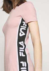Różowa dopasowana sukienka z krótkimi rękawami, posiadająca czarno-białą pionową pasek boczny z wyboldowanym napisem "FILA". Gładka tekstura.