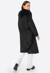 Černý vlněný kabát s velkou kapucí, uvázaným pasem a měkkou texturou. Nositeli ho doplňují bílé kalhoty a robustní černé boty.
