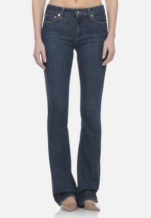 Donna che indossa jeans bootcut blu scuro e scarpe beige con punta a punta, in piedi contro uno sfondo bianco.